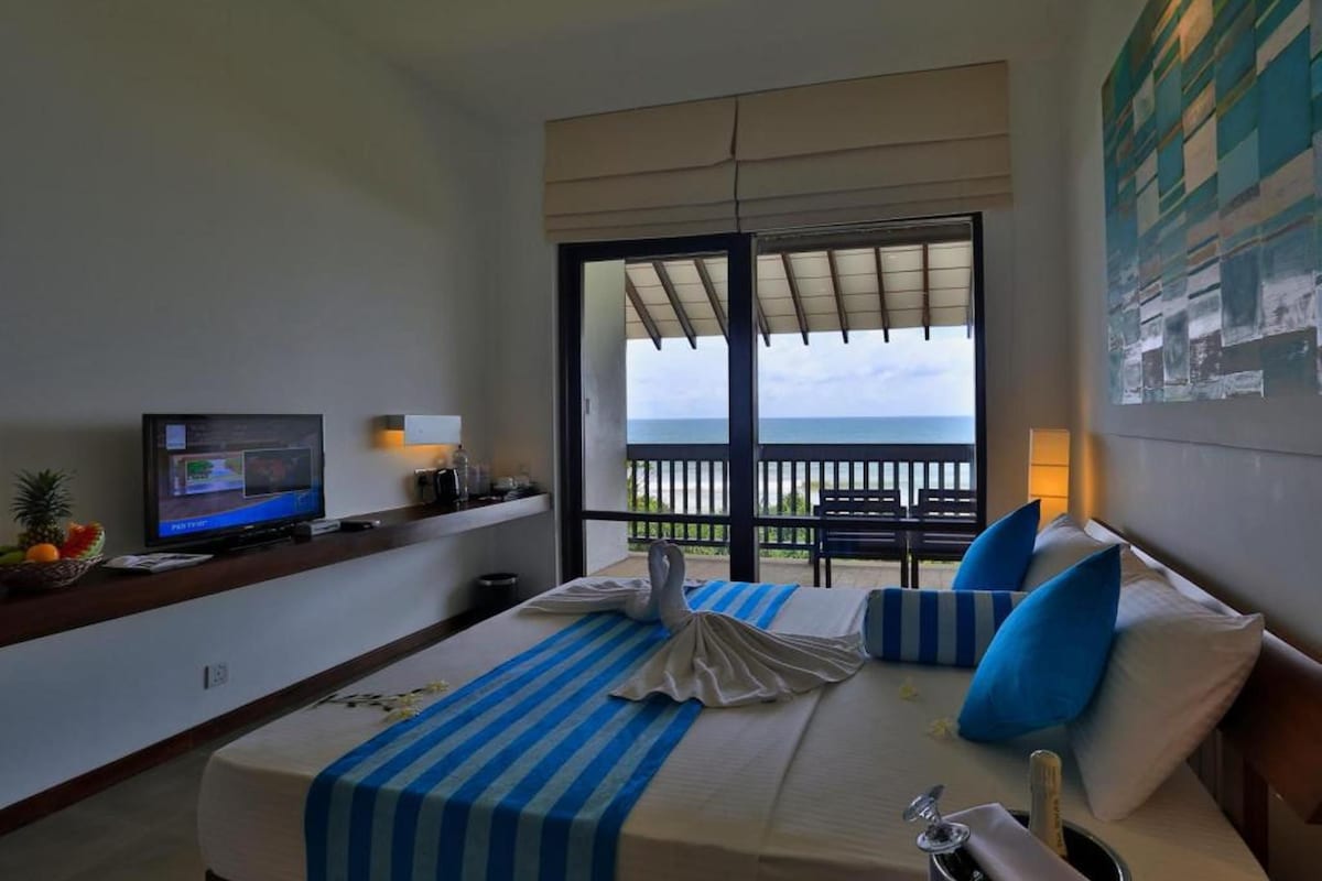 A hotel room with a bed, a television, a balcony with a view of the ocean, and a painting on the wall.