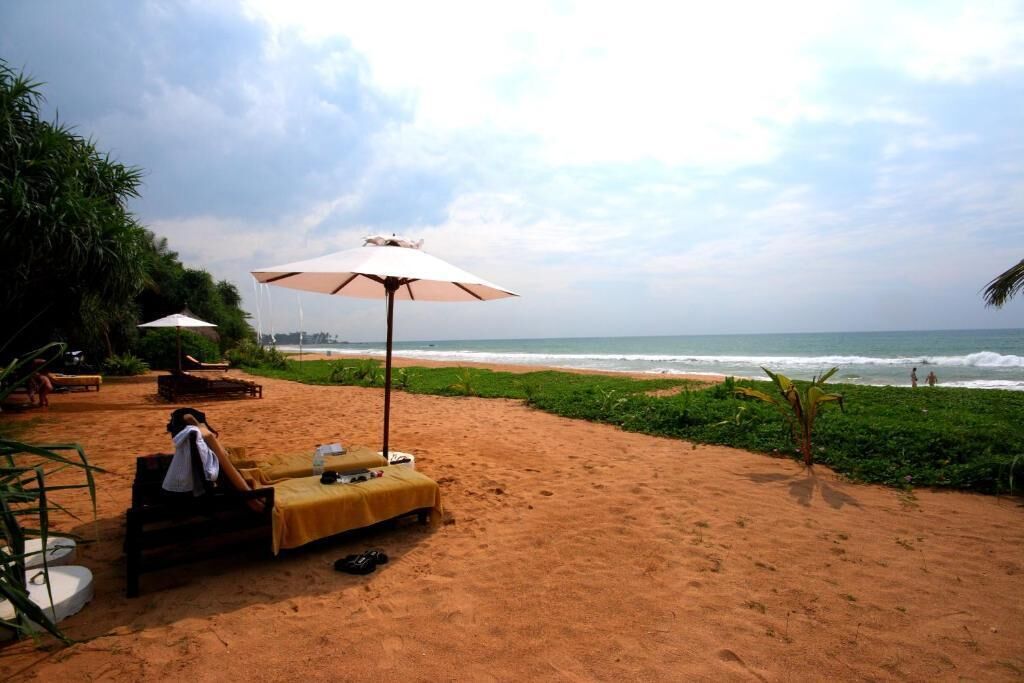 A beachside setting with a person relaxing under an umbrella, a table with items, and a view of the ocean.