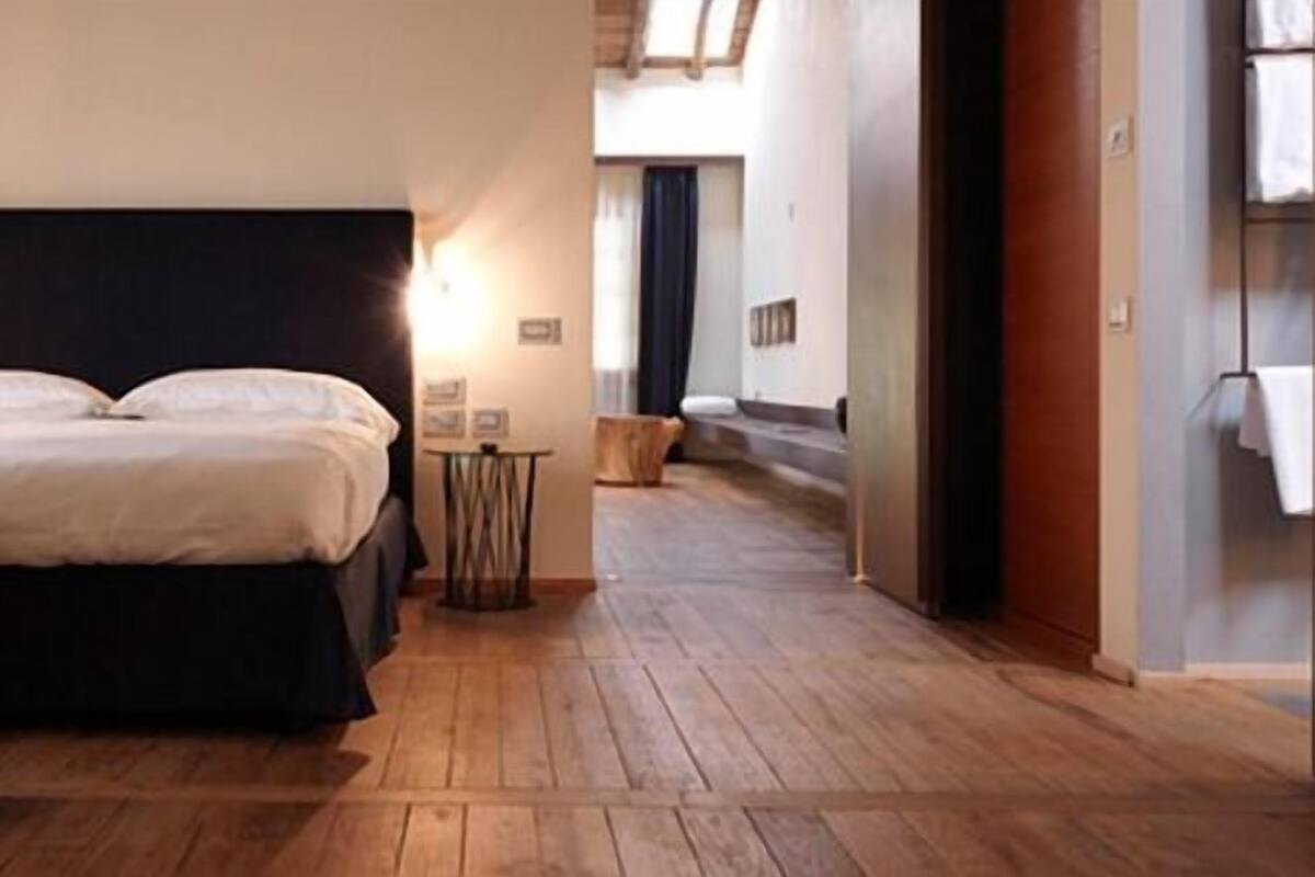 A bedroom with a large bed, wooden flooring, and a nightstand with a lamp.