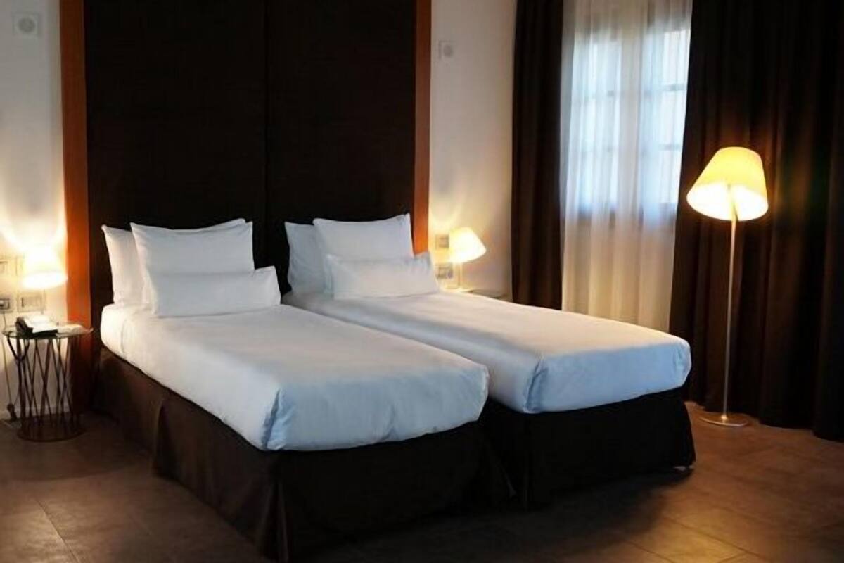 A hotel room with two single beds, a dark headboard, a bedside table with a lamp, and a window with curtains.