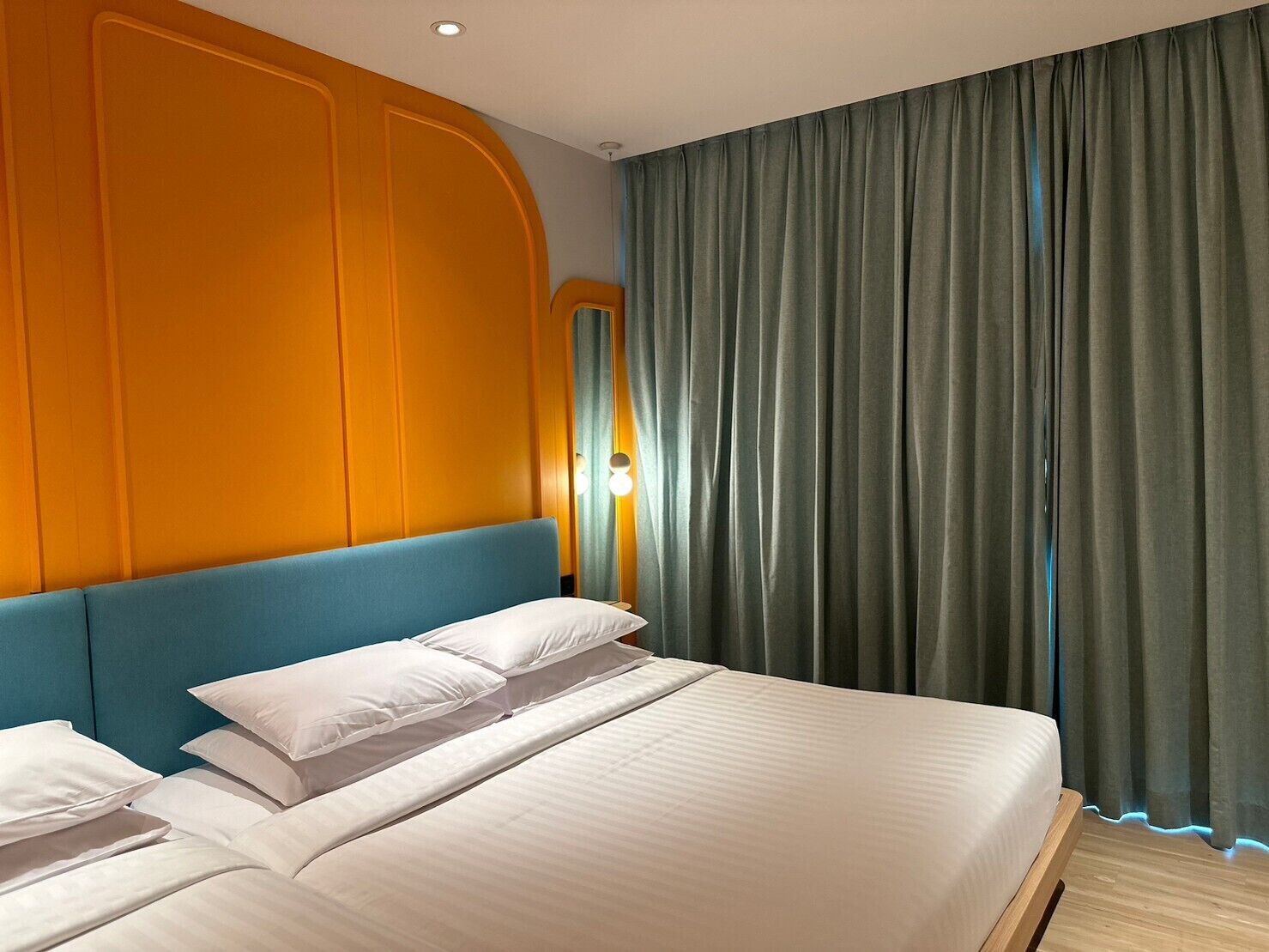 Habitación de hotel con una cama grande, paneles de pared naranja y vistas de la ciudad a través de cortinas verdes.