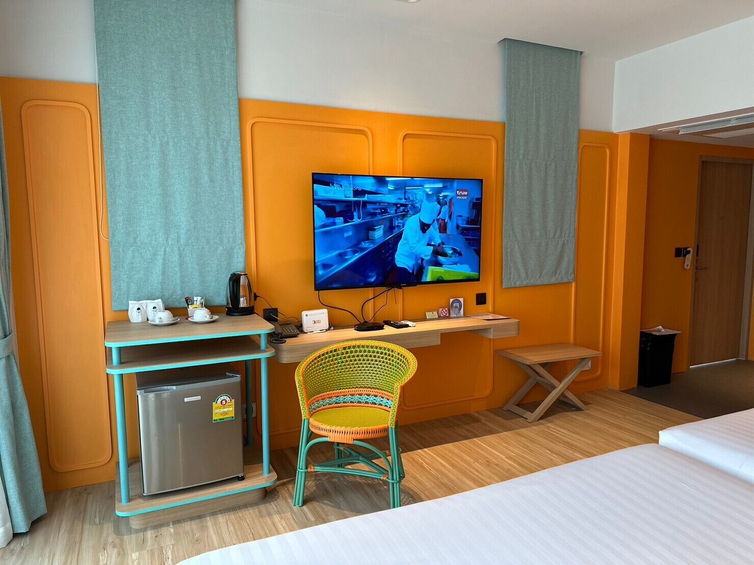 A hotel room with a TV mounted on the wall, a small kitchenette, a wicker chair, and a bed.