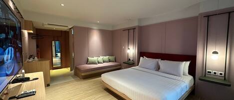 A modern hotel room with a large bed, a sofa, a desk with a TV, and a small bedside table.