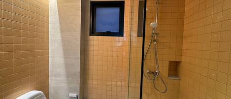 Un baño pequeño con inodoro, un bote de basura, ducha con cabezal de ducha manual y una ventana sobre el lavamanos.