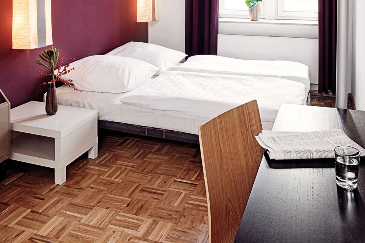 A hotel room with a bed, a nightstand, a table with a glass of water, and a window with curtains.