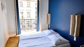 Una habitación con piso de madera, una cama con ropa blanca, una ventana con cortinas y una pared azul con un libro colgado.