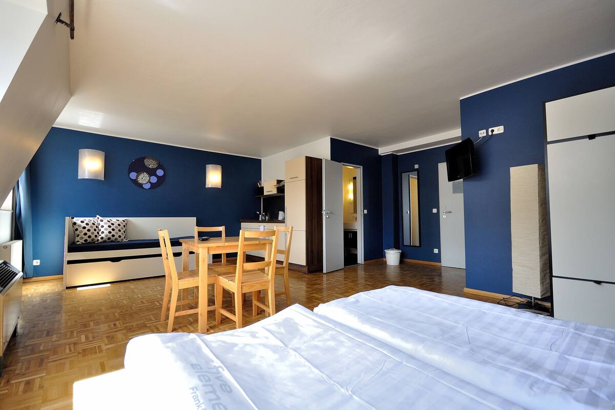 A compact room with a bed, a small table, and chairs. There is a kitchenette with a fridge and a microwave. The room has blue walls and a wooden floor.