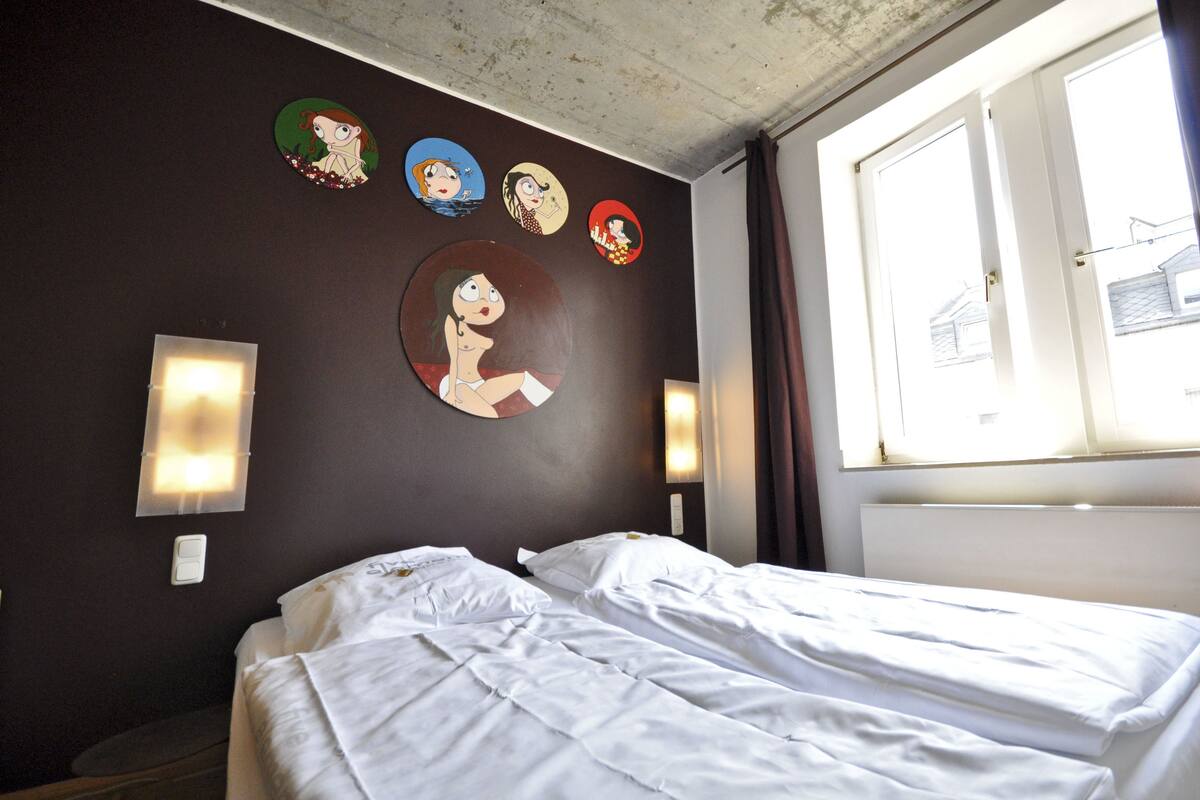 A hotel room with two beds, a window with curtains, wall-mounted light fixtures, and cartoon character decorations.