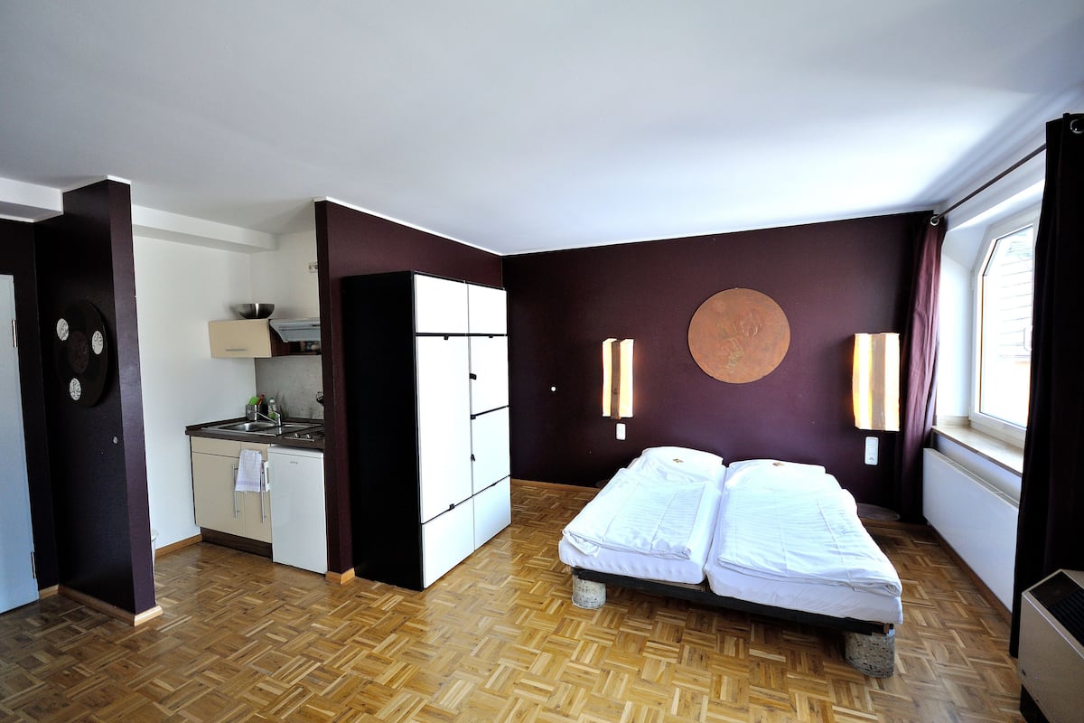 A compact room with a bed, a kitchenette, and a large window.