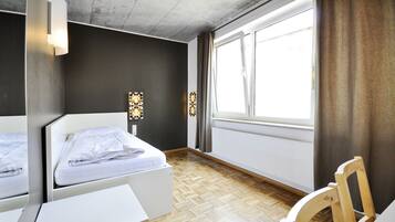 Une chambre d’hôtel comprenant un lit, un bureau, une chaise, une fenêtre avec des rideaux et un luminaire fixé au mur.