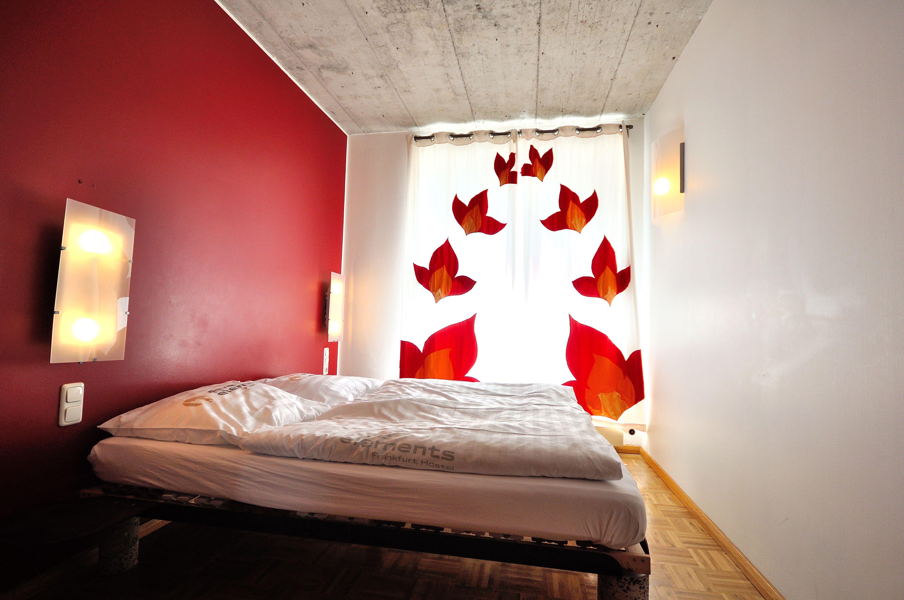 Una habitación con cama, pared roja y ventana con cortina de diseño de hojas rojas.