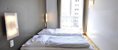 A hotel room with a bed, a light switch, and a view of a building through a window.