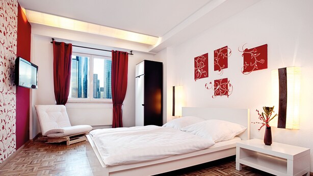 A modern bedroom with a bed, a bedside table, a chair, and a TV mounted on the wall. - Five Elements Hostel (Frankfurt)