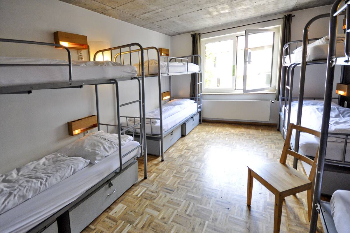 A dormitory room with bunk beds, a wooden chair, and a window providing natural light.