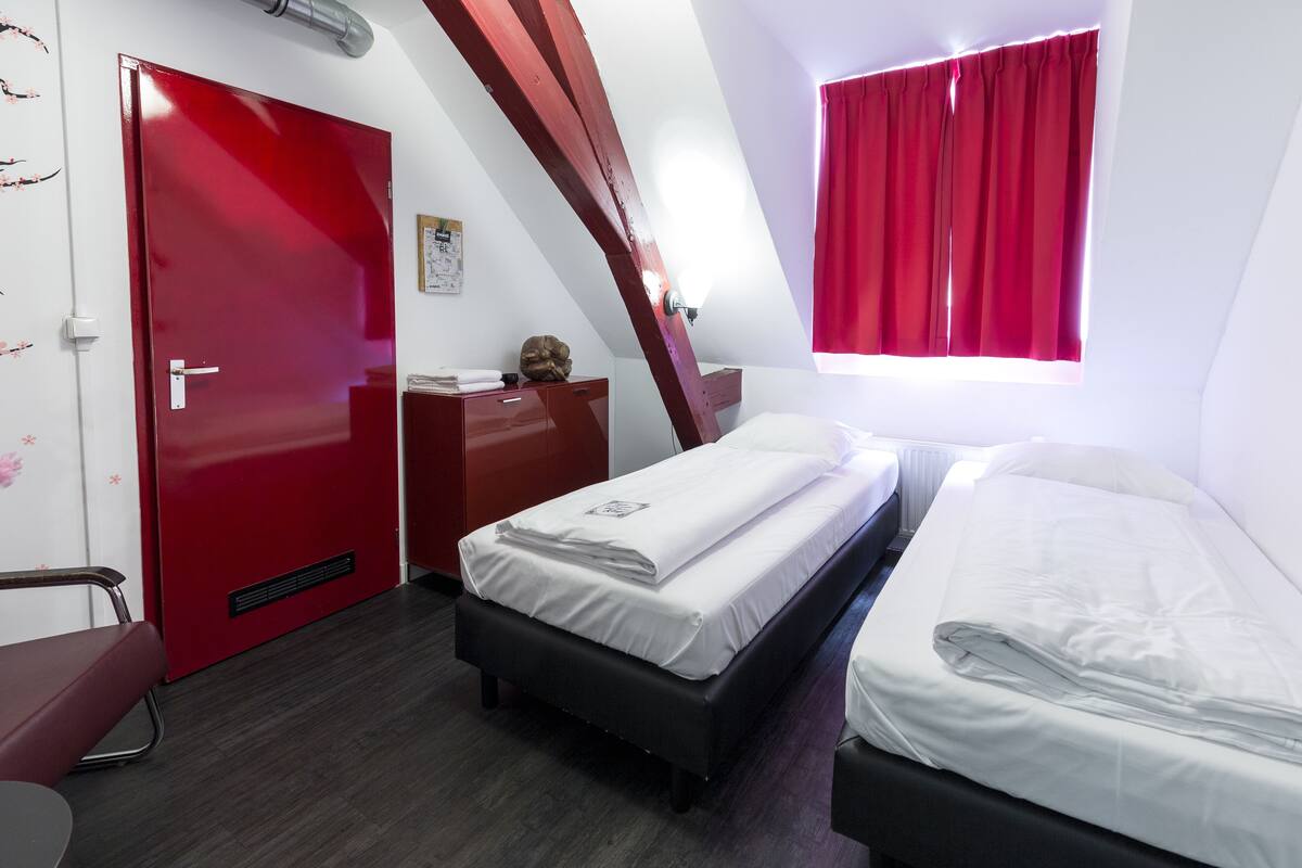 A compact hotel room with two single beds, a red door, a small desk, and a window with red curtains.