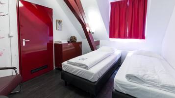 A compact hotel room with two single beds, a red door, a small desk, and a window with red curtains.