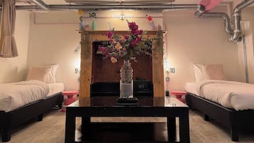 A hotel room with two beds, a central table, a fireplace with a vase of flowers, and visible numbers on the walls.