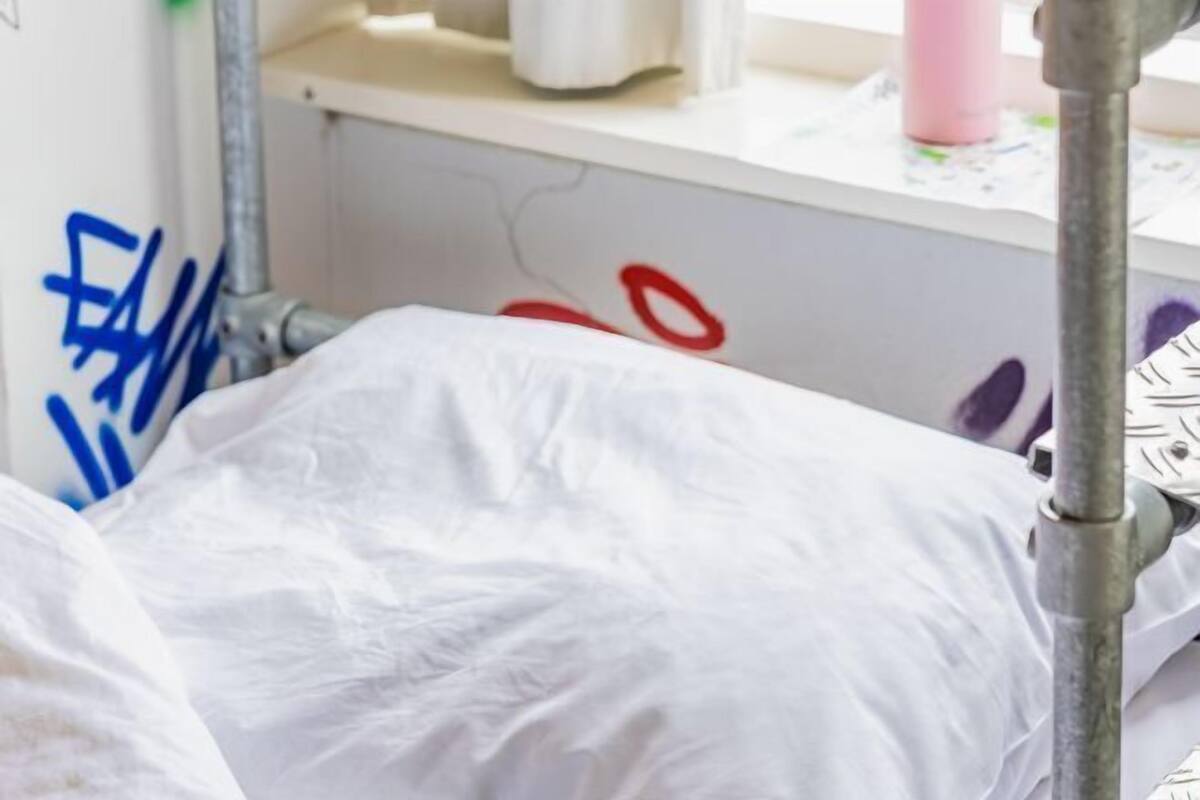 A hospital bed with white sheets, a metal pole, and a wall with graffiti.