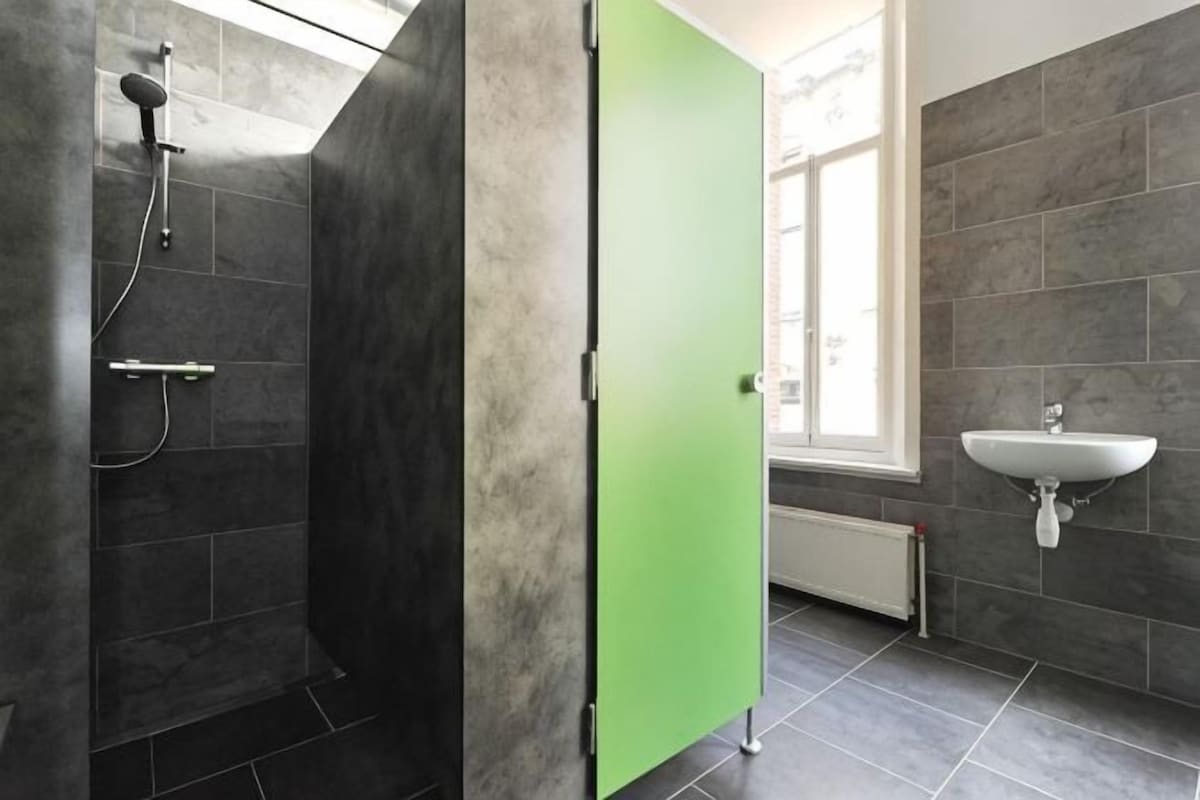 Uma casa de banho com chuveiro, porta verde, pia e janela.
