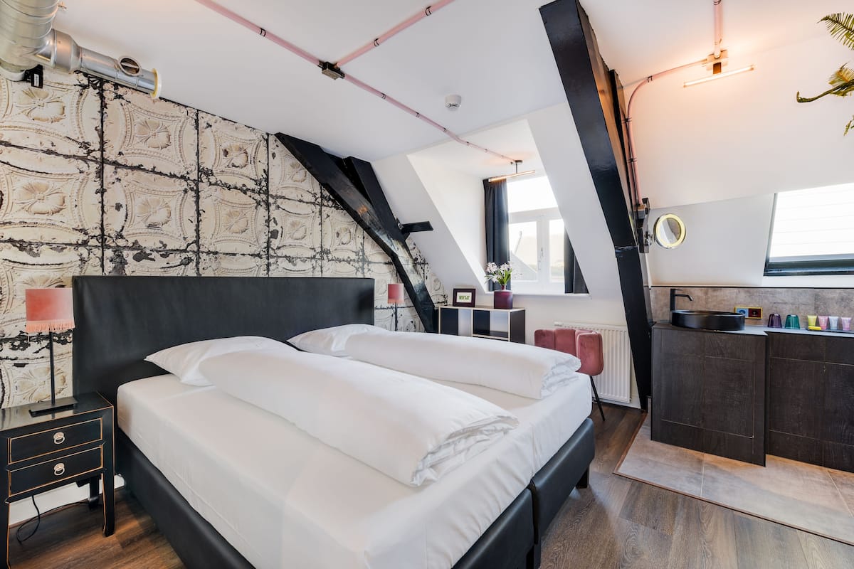 A bedroom with a large bed, a small desk, a nightstand, a window, and a kitchenette.