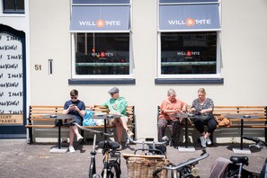 Three people seated at tables outside a Will & Tate coffee shop, with bicycles and a sign on the wall. - Will & Tate City Stay (The Hague)