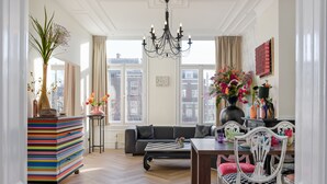 A living room with a sofa, a dining table, and chairs. There is a chandelier, a vase with flowers, and a bookshelf. - Will & Tate City Stay (The Hague)