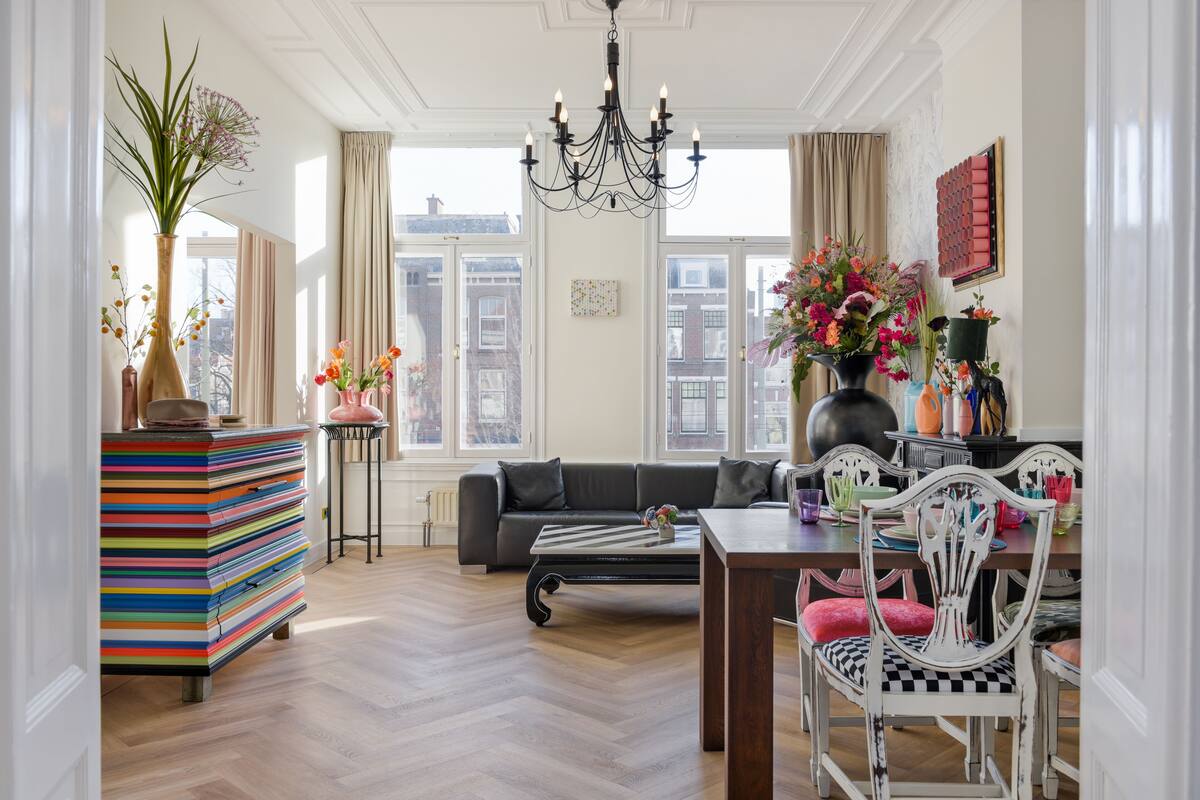 A living room with a sofa, a dining table, and chairs. There is a chandelier, a vase with flowers, and a bookshelf.
