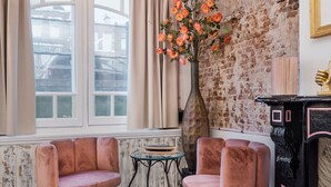 A room with a brick wall, two pink chairs, a small table, a vase with flowers, and a black cabinet with pink trim. - Will & Tate City Stay (The Hague)