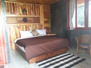 A wooden cabin bedroom with a bed, pillows, a nightstand, a wicker chair, and a window with curtains.
