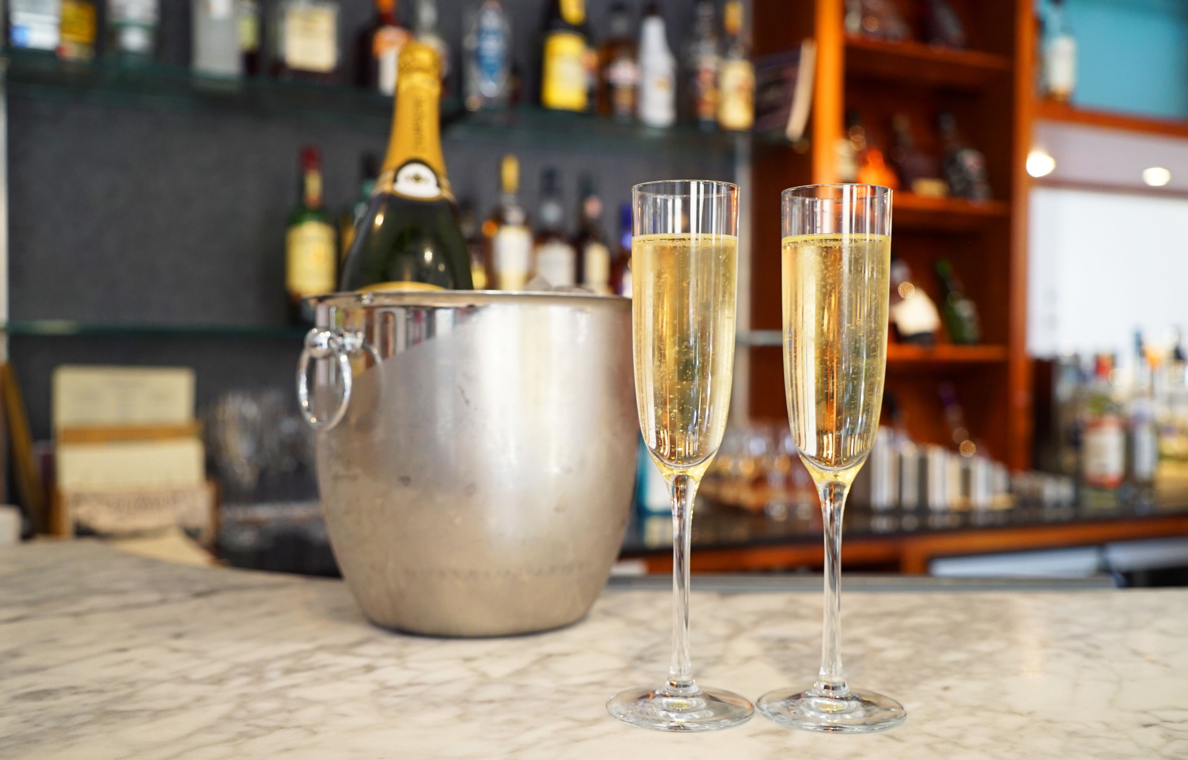 Two champagne flutes on a marble countertop with a large metal bucket in the background.