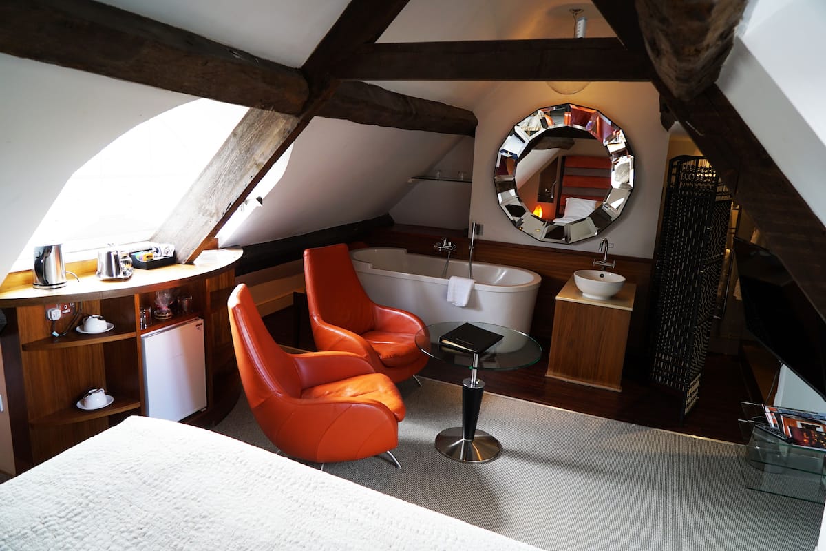 A modern attic bedroom with a sloped ceiling, a round mirror, a bathtub, a red leather chair, a small table, and a white bed.