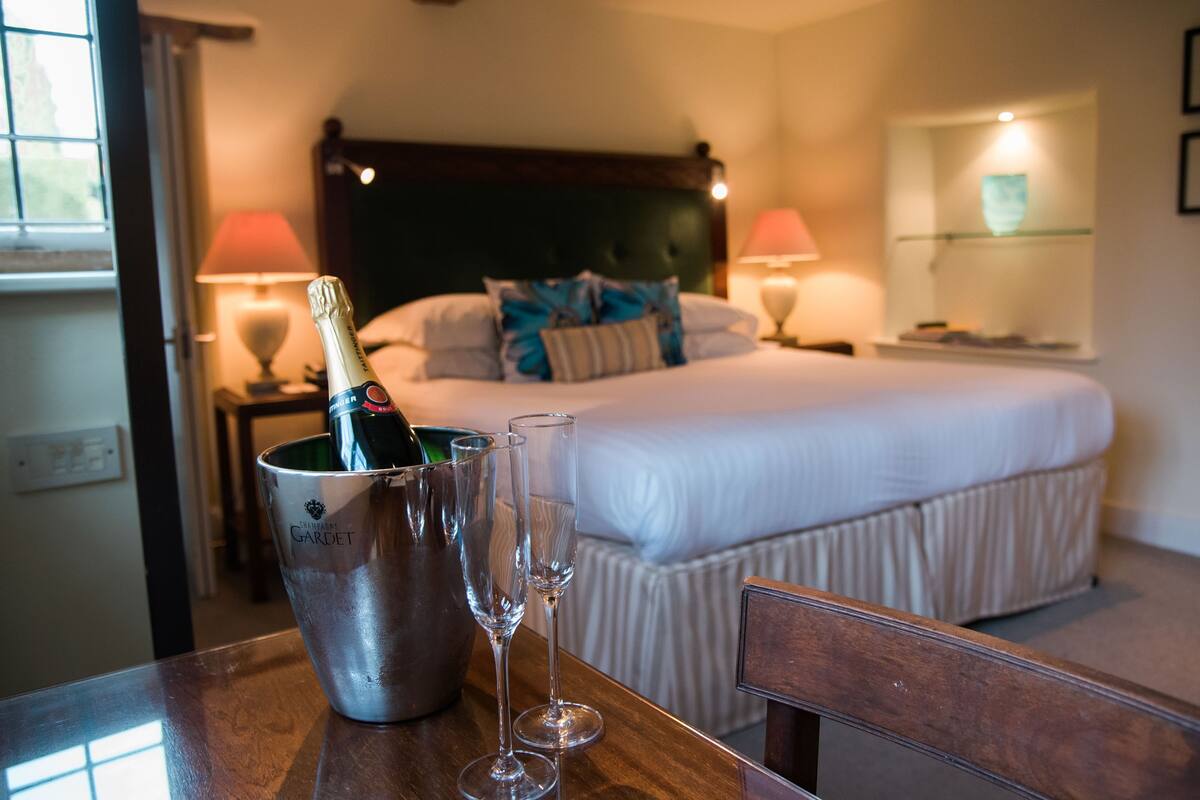 A hotel room with a bed, bedside lamps, a nightstand, a bottle of champagne, and two champagne flutes on a table.