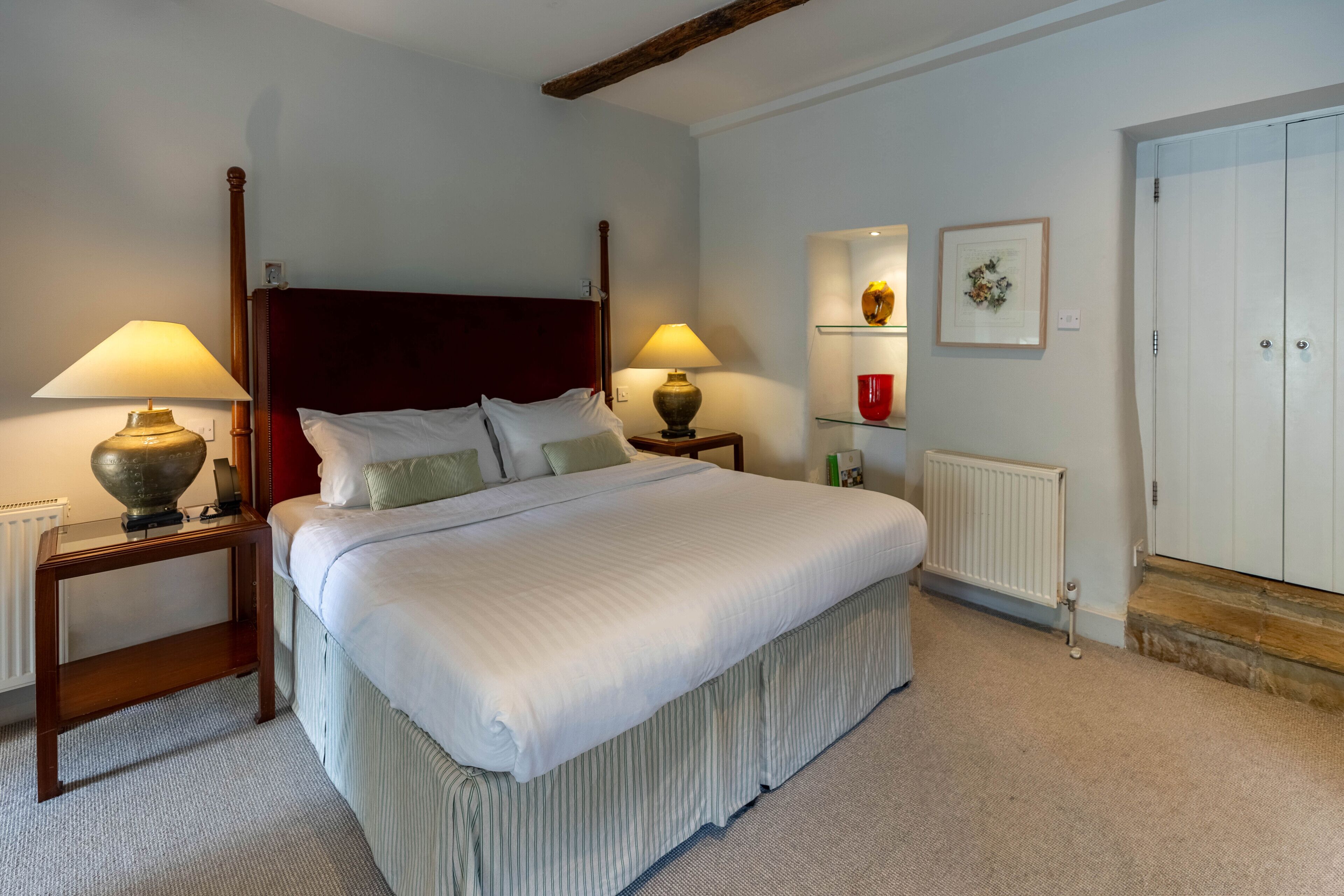 A neatly arranged bedroom with a large bed, two bedside tables with lamps, a radiator, a white wardrobe, and a shelf with decorative items.