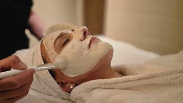A person lying on a bed covered with a white towel, receiving a facial treatment.