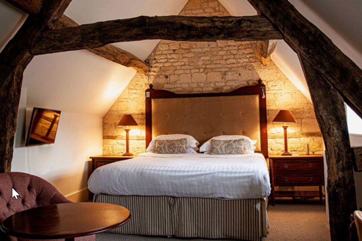 A bedroom with a stone wall, a wooden ceiling, a bed with white bedding, two bedside tables with lamps, and a chair.