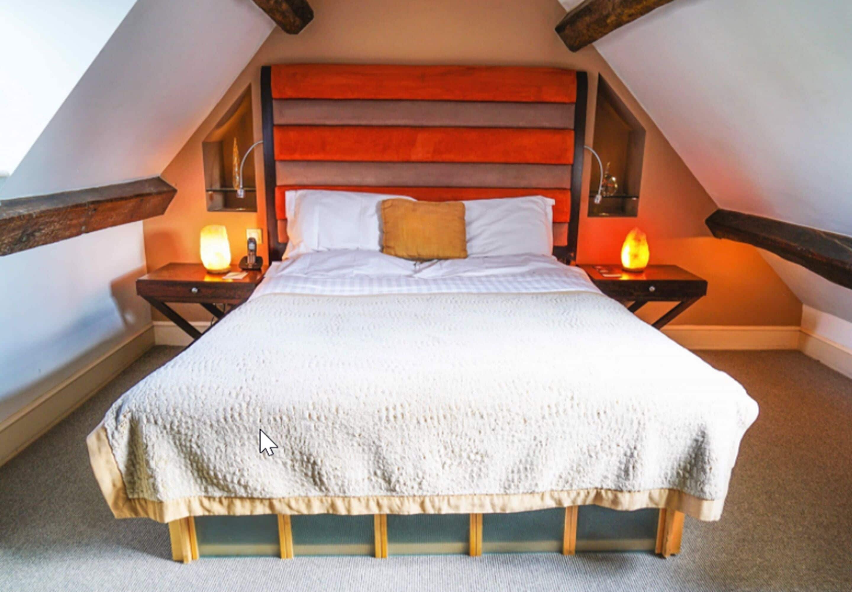 Een slaapkamer met een schuine kap, een groot bed met een witte dekbedovertrek, twee nachtkastjes met lampen, en een houten hoofdbord.