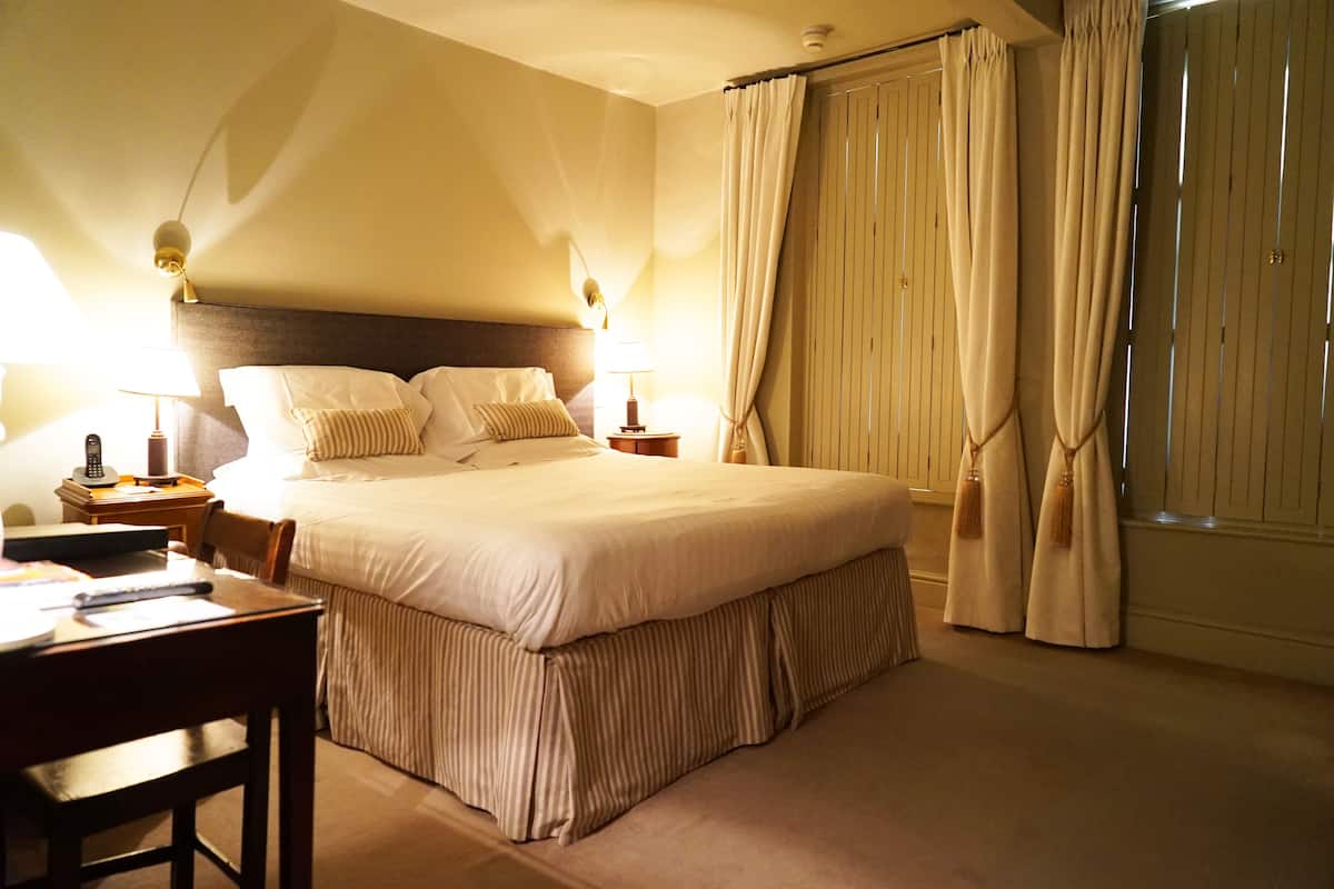 A neatly made bed with a headboard, two bedside tables with lamps, a wooden chair, and a window with curtains.