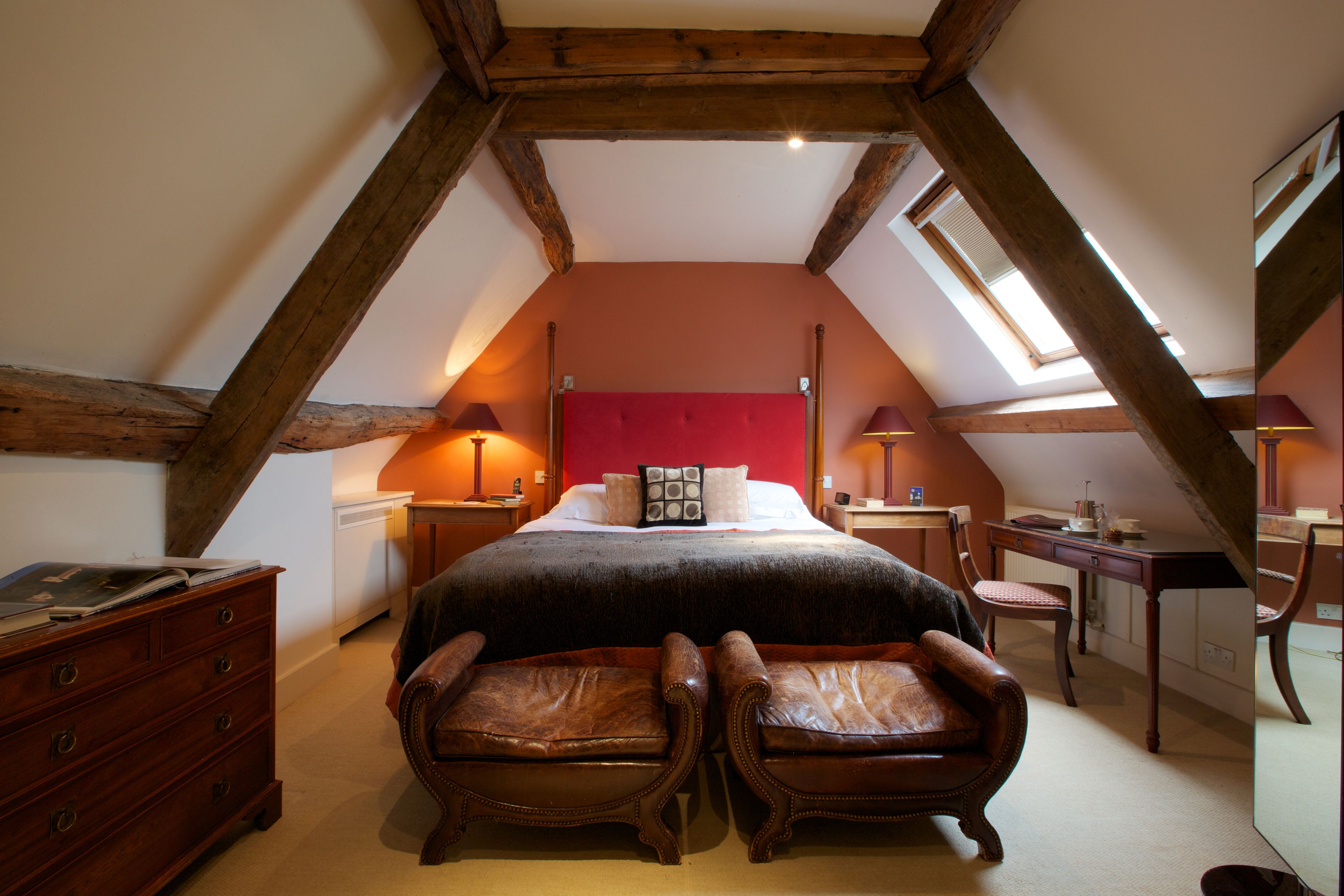Une chambre avec un plafond en pente, un lit avec une tête de lit rouge, deux fauteuils en cuir, une commode et un bureau avec une lampe.