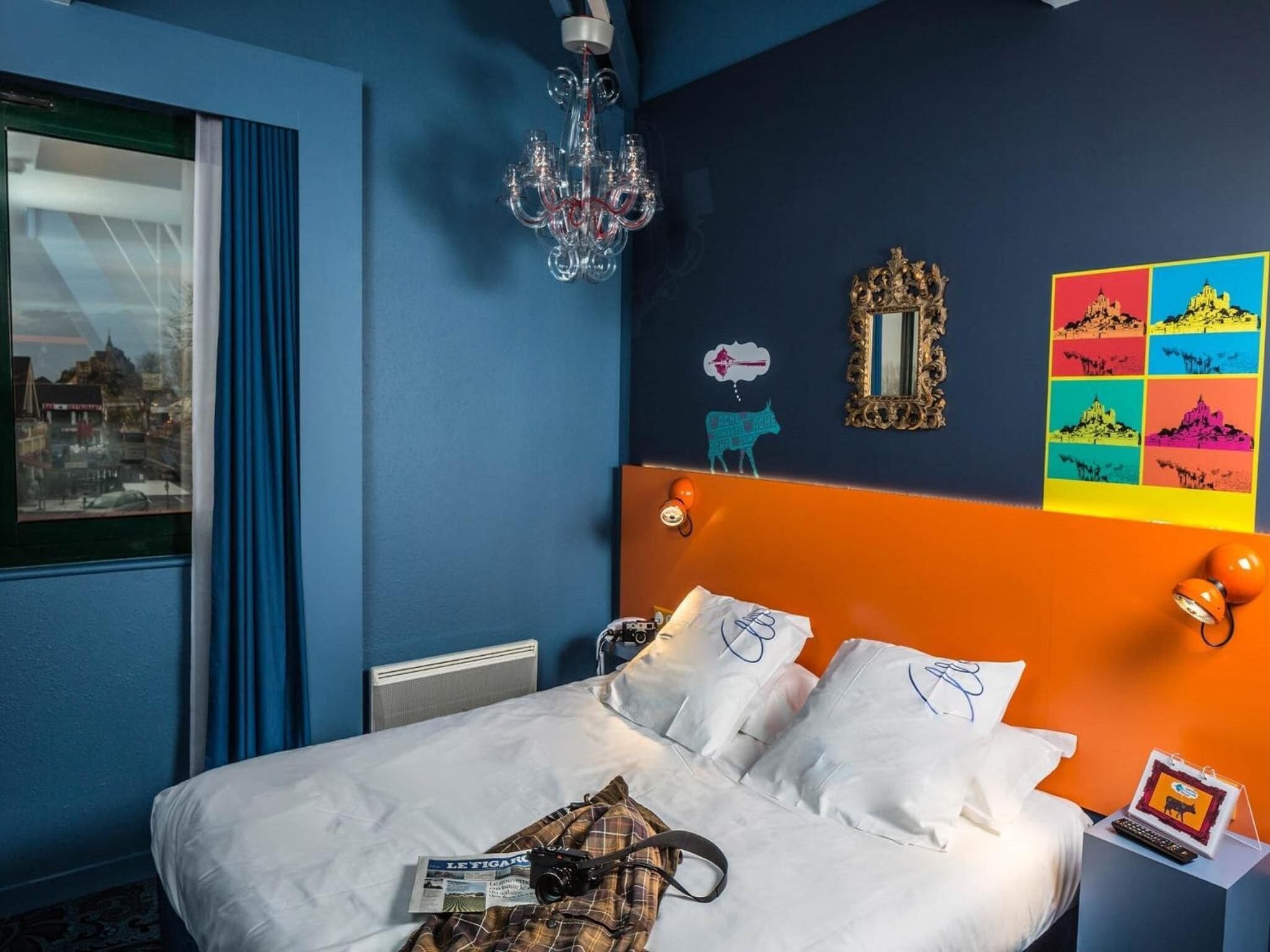 A bedroom with a bed, a chandelier, a window with a view, and a colorful wall decoration.