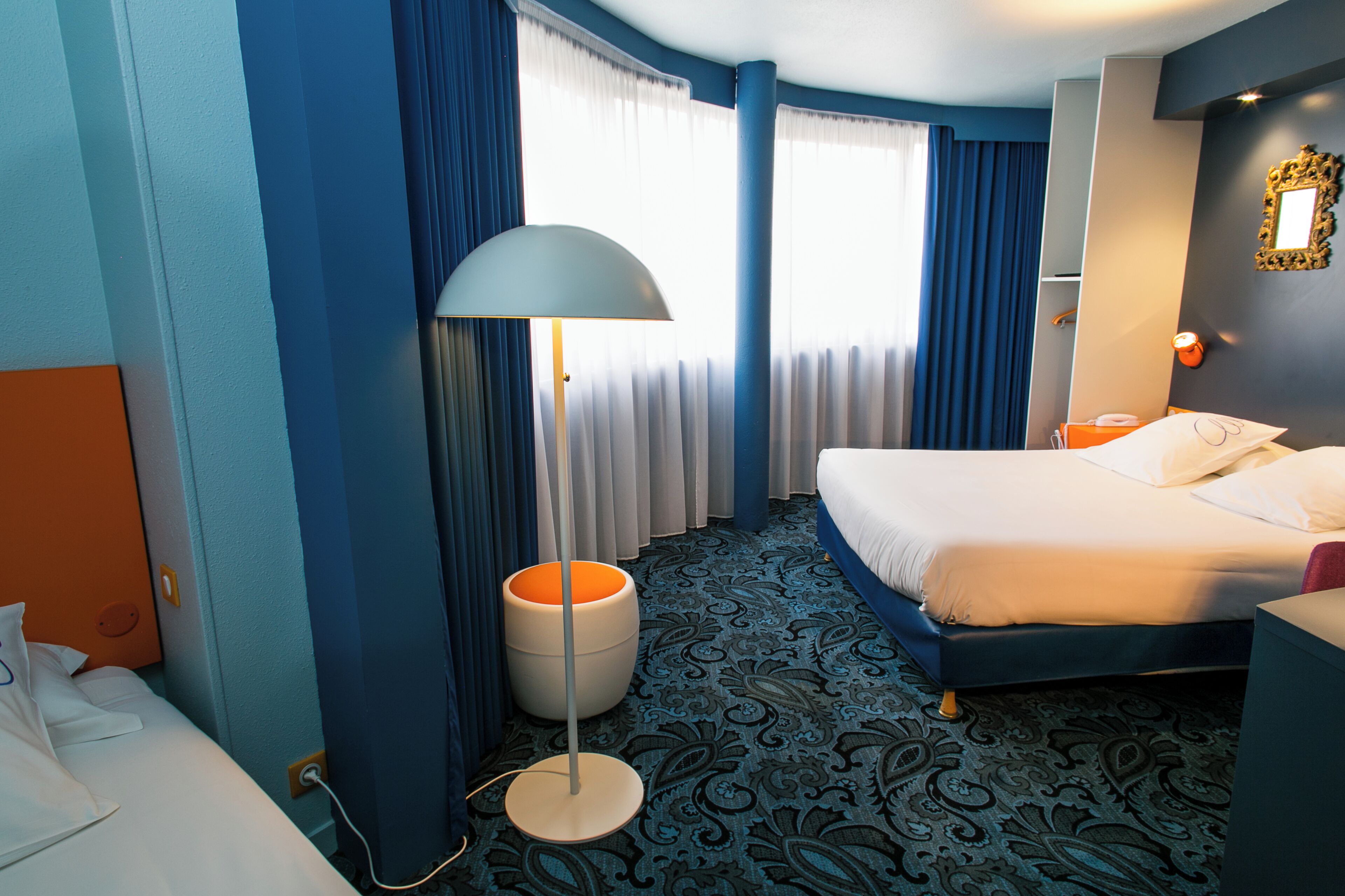 A hotel room with a bed, a floor lamp, a trash bin, and a window with curtains.