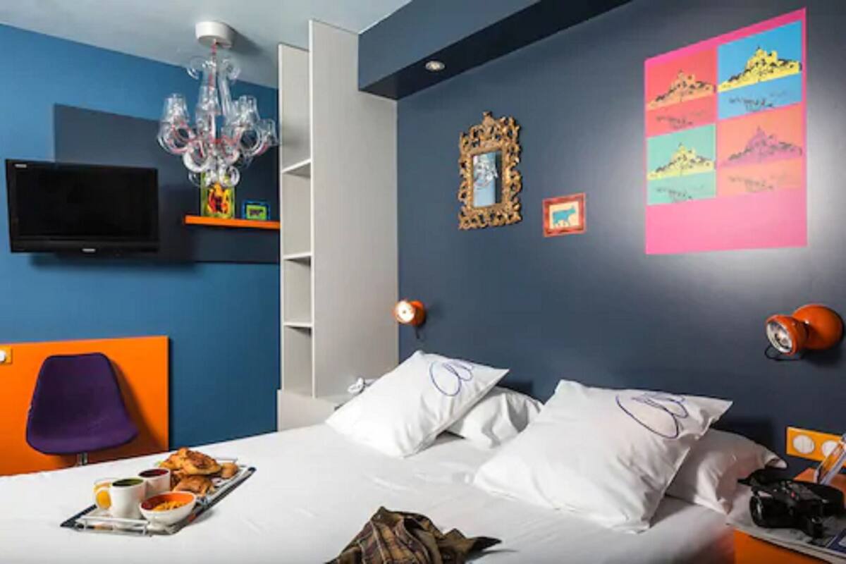 Ein Hotelzimmer mit Bett, Kissen, einem Tablett mit Speisen, einem Fernseher, einer Chandelierleuchtre und Wanddekorationen.