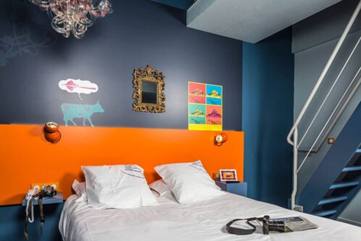 Ein Hotelzimmer mit Bett, Nachttisch, Wand mit dekorativem Spiegel, farbenfroher Kunst und einer Treppe.