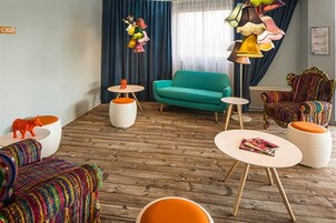 Una sala moderna con un sofá verde azulado, sillones coloridos y piso de madera. Hay mesitas redondas y una lámpara colorida.