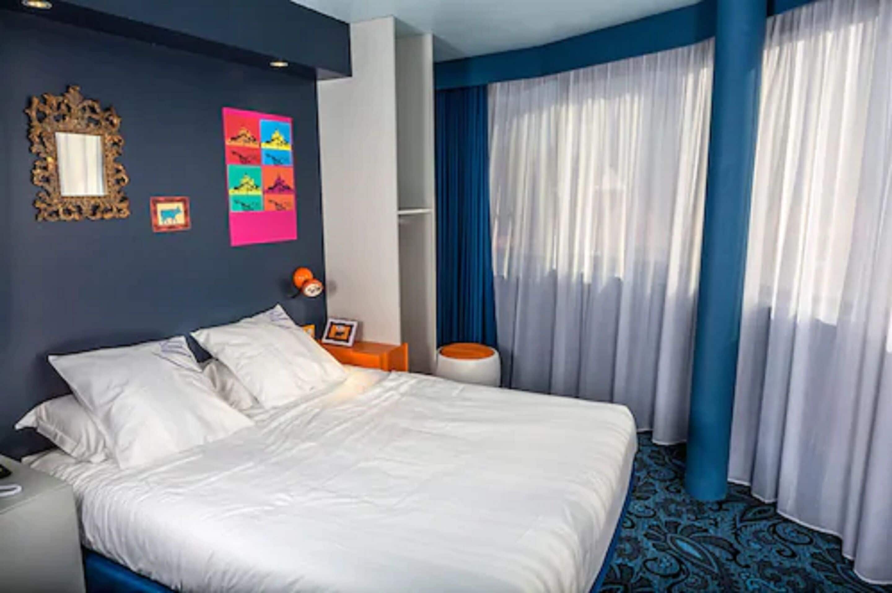 Habitación de hotel con una cama grande, una mesita de noche, un espejo y una ventana con cortinas azules.