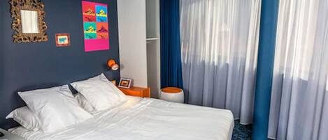 Una camera d'albergo con un letto grande, un comodino, uno specchio e una finestra con tende azzurre.