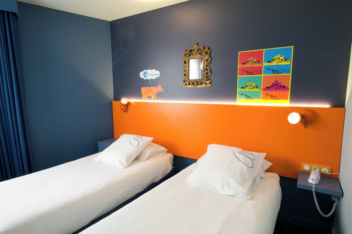 Ein Hotelzimmer mit zwei Einzelbetten, einem Nachttisch mit Telefon sowie einer Wand mit Spiegel und farbenfroher Kunst.