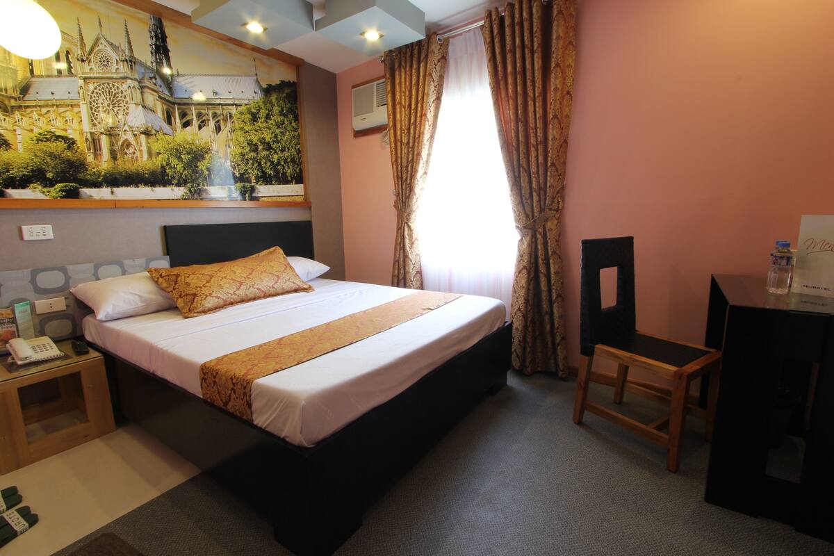 A hotel room with a large bed, a desk, a chair, a nightstand, and a wall-mounted air conditioner.