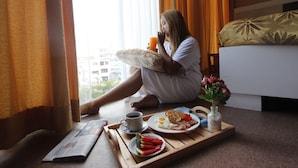 A person sitting by a window with curtains, holding a cup and a pillow, with breakfast items on a tray.