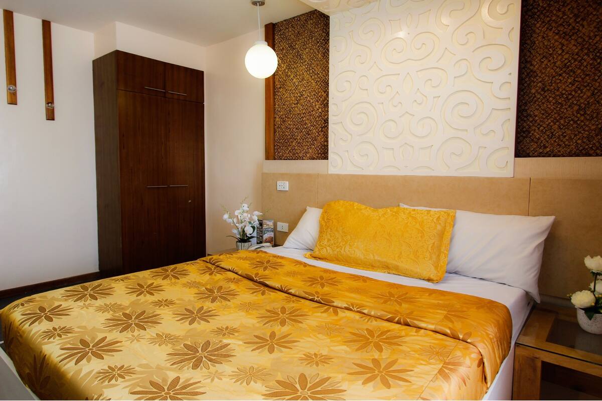 A hotel room with a bed, a wooden wardrobe, a decorative wall panel, and a small table with a vase of flowers.