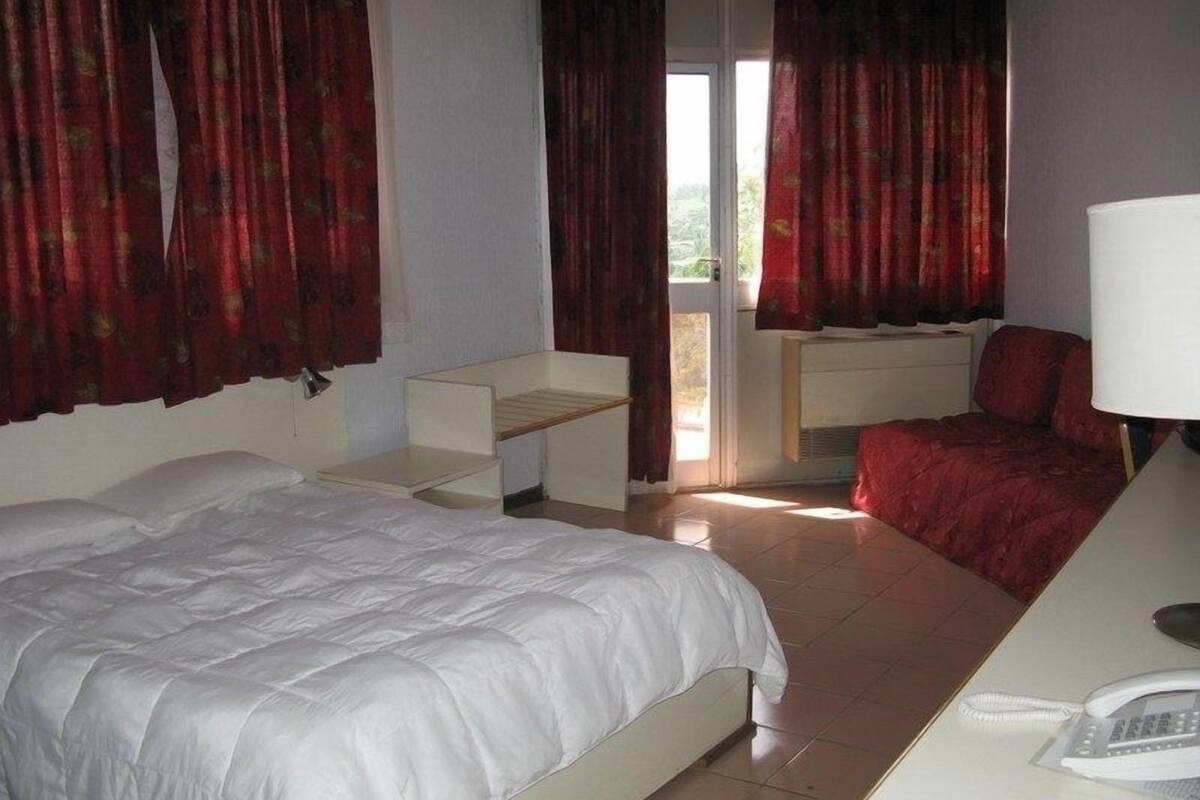 A bedroom with a bed, a desk with a lamp, a chair, and a window with curtains.